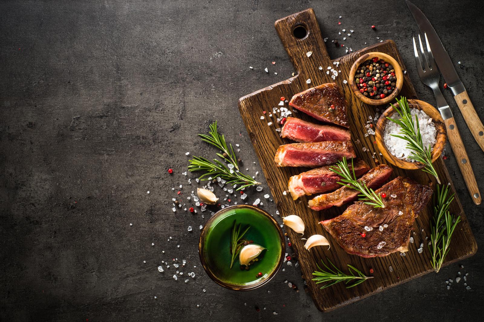 Steak on a wooden cutting board with herbs and spices on a dark surface